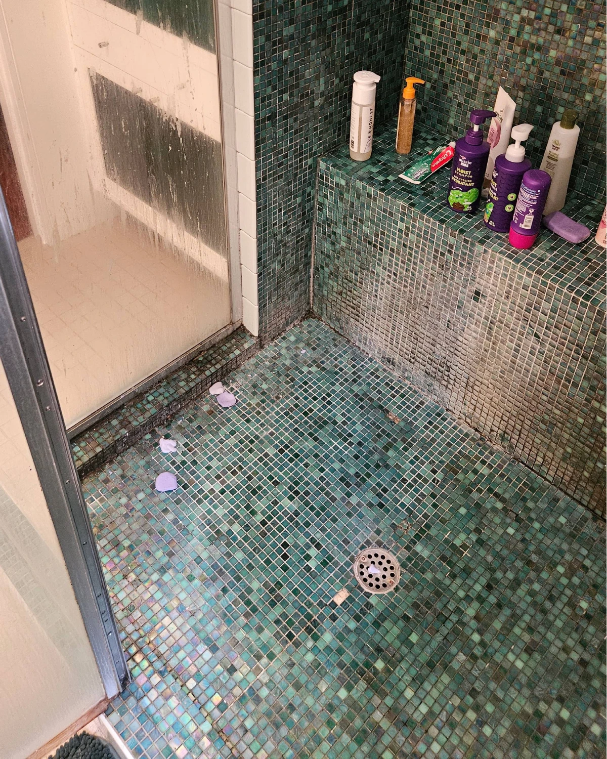 Shower glass, green mosaic floor, and tiled bench covered with soap scum before deep cleaning